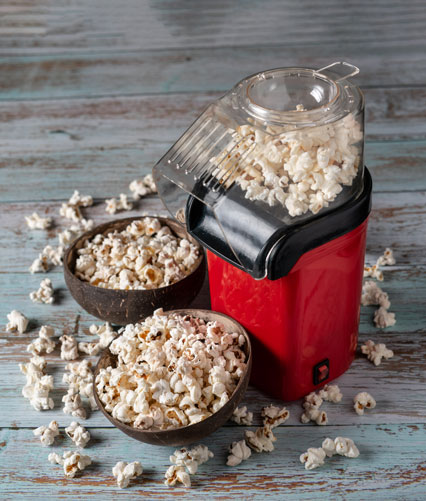 red popcorn popper and two bowls of popcorn on wooden surface. Additional popcorn is scattered about.